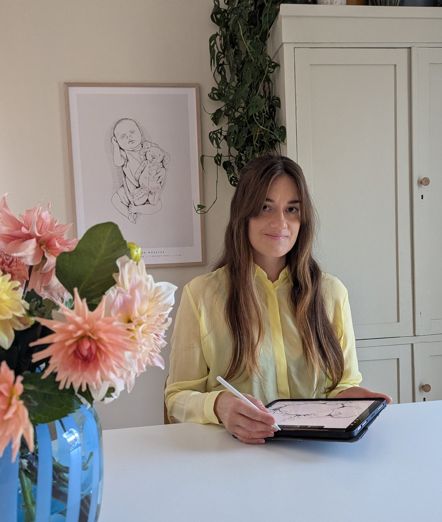 Artist sitting at a desk drawing, with a hand-drawn birth poster hanging on the wall in the background